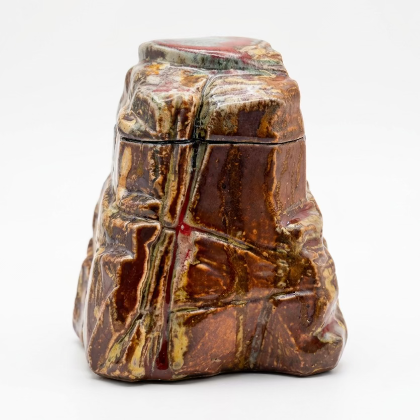 A handmade ceramic lidded container or vase with a highly textured, sculptural design. The piece features a glossy, earthy brown and reddish-brown glaze with prominent metallic gold and green veins running through the surface, set against a neutral light gray backdrop.
